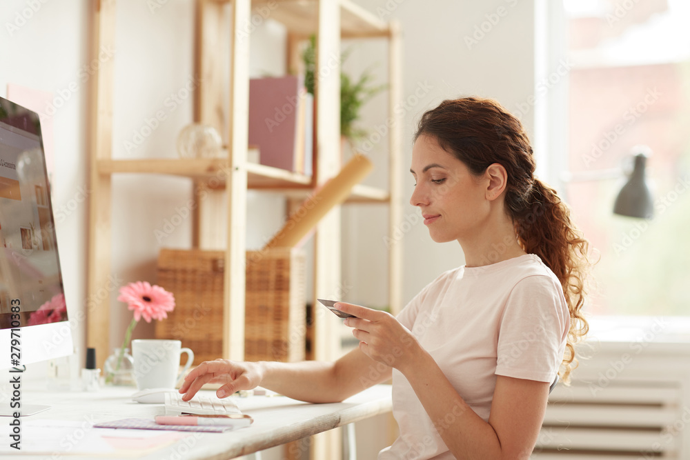 Serious pensive young lady with pony tail sitting at table and typing on keyboard while filling card details on website Serious pensive young lady with pony tail sitting at table and typing on keyboard while filling card details on website