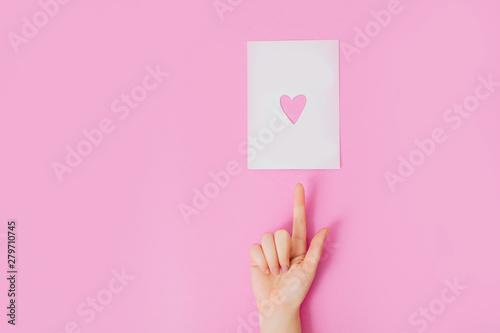 Female hand with manicure on a pink background pointing at a white sheet of paper with heart shaped hole in it.