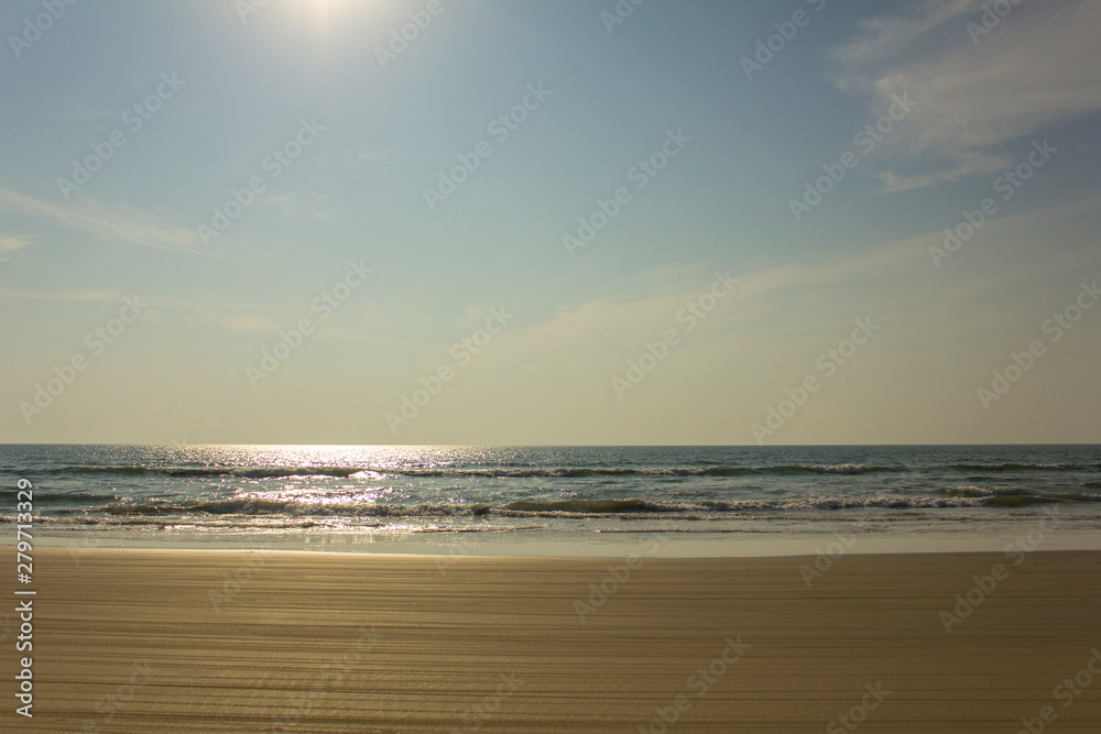 yellow sandy beach with motorcycle tire tracks against the backdrop of sea waves under a clear blue sky with the sun