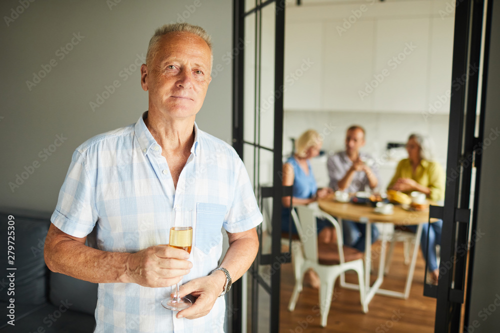 Obraz premium Waist up portrait of handsome mature man holding champagne flute looking at camera posing at home with friends in background, copy space
