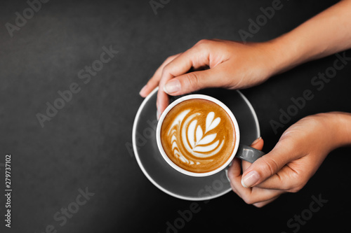 Cup of fresh cappuccino in woman hands on dark black metal table background. Empty place for text. Coffee addiction. View from above in hipster cafe. photo