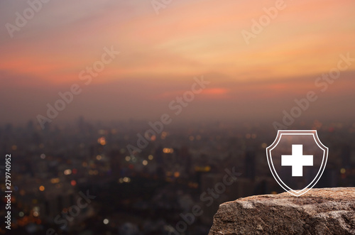 Cross shape with shield flat icon on rock mountain over blur of cityscape on warm light sundown, Business security insurance concept