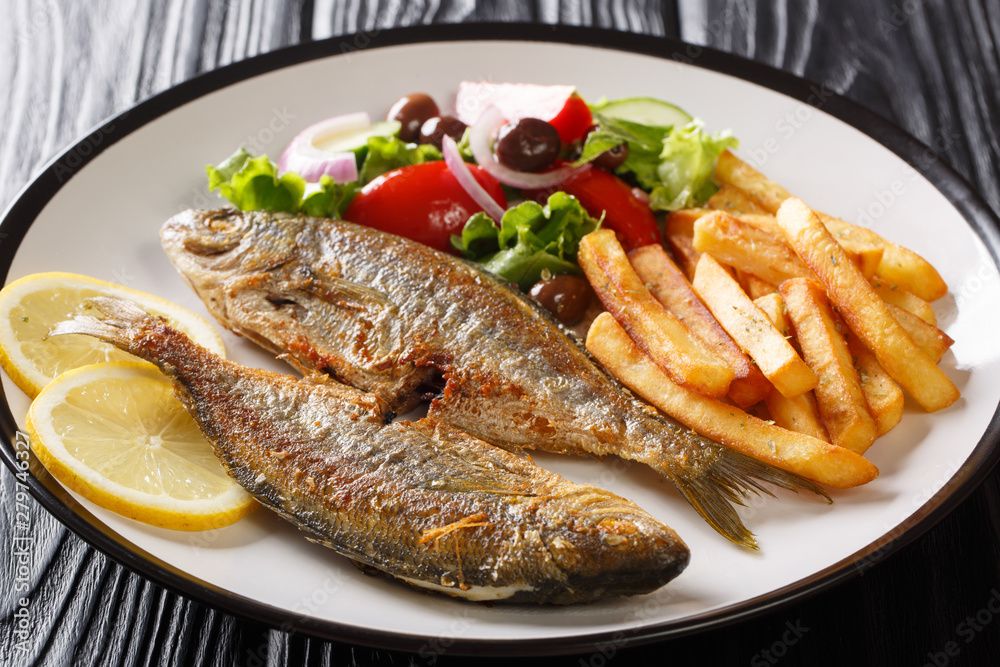 Mediterranean cuisine Fried Sarpa fish with fresh vegetable salad, lemon and french fries close-up on a plate. horizontal Mediterranean cuisine Fried Sarpa fish with fresh vegetable salad, lemon and french fries close-up on a plate. horizontal