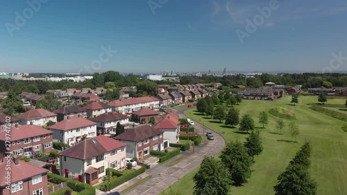 An ascending shot looking out over trees and grass in an urban green space on a Summer day. You can see many of the houses in the area with Manchester skyline in the background.