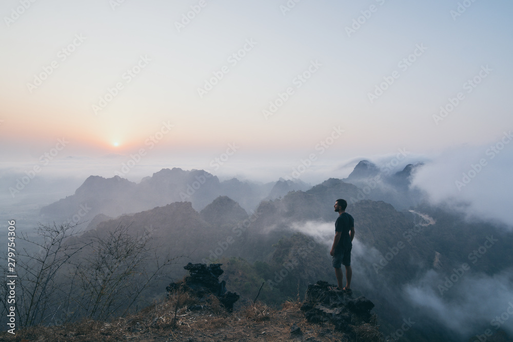 Man standing on the rock overlooking tropical mountains at sunrise foggy morning in Hpa-an, Myanmar