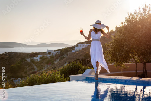 Elegante Frau in weißem Sommerkleid steht am Pool und genießt den Sonnenuntergang mit einem Cocktail in der Hand photo