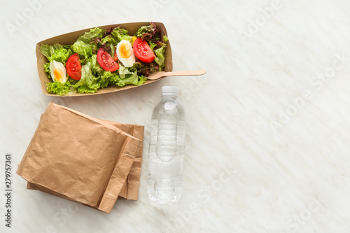 Bottle of water, paper box and bags with delicious food on light background
