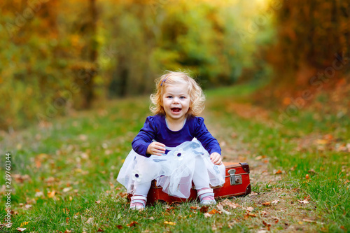 Cute little toddler girl sitting on suitcase in autumn park. Happy healthy baby enjoying walking with parents. Sunny warm fall day with child. Active leisure and activity with kids in nature. © Irina Schmidt