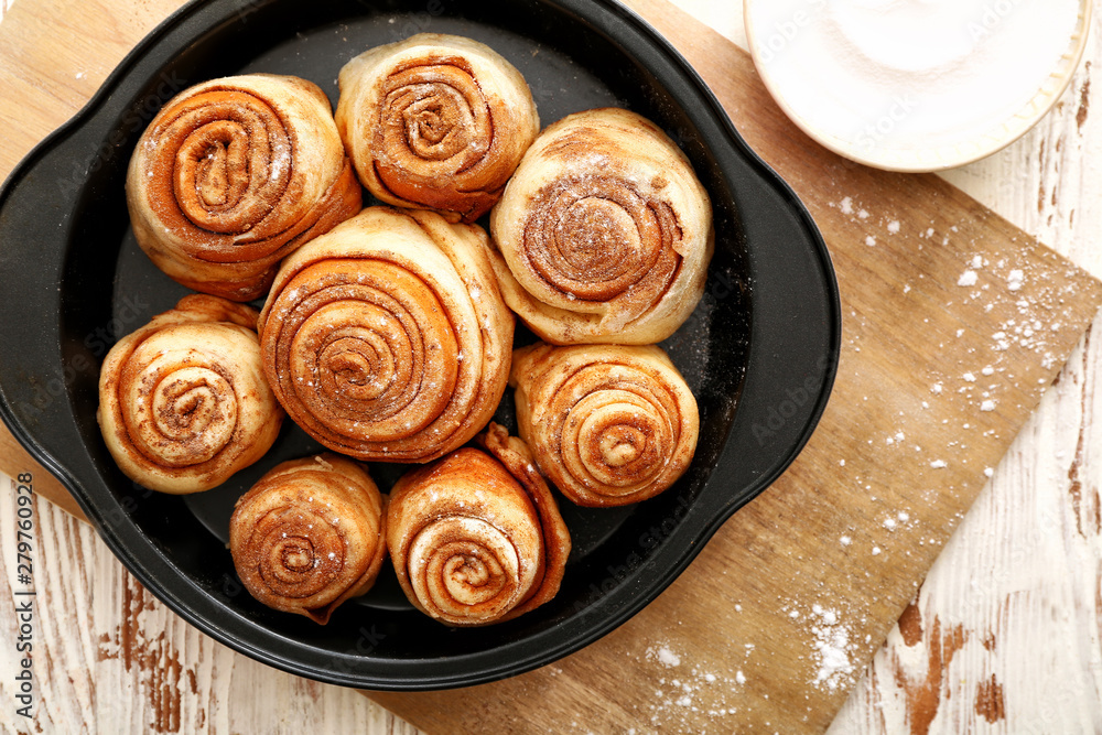 Fototapeta premium Freshly baked cinnamon rolls in black baking pan with powdered sugar on wooden board, overhead view