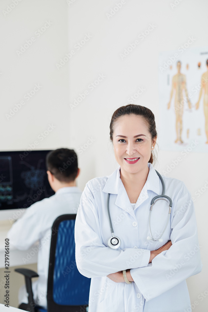 Smiling pretty Asian mature doctor in labcoat folding arms and looking at camera Smiling pretty Asian mature doctor in labcoat folding arms and looking at camera