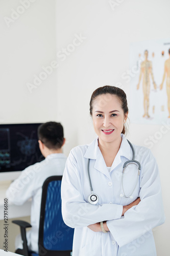Smiling pretty Asian mature doctor in labcoat folding arms and looking at camera