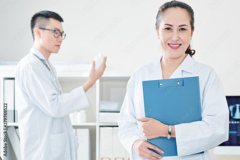 Cheerful mature Vietnamese doctor with clipboard smiling and looking at camera Cheerful mature Vietnamese doctor with clipboard smiling and looking at camera