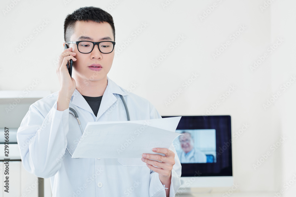 Young serious Vietnamese doctor reading document, talking on phone and telling diagnosis to client Young serious Vietnamese doctor reading document, talking on phone and telling diagnosis to client