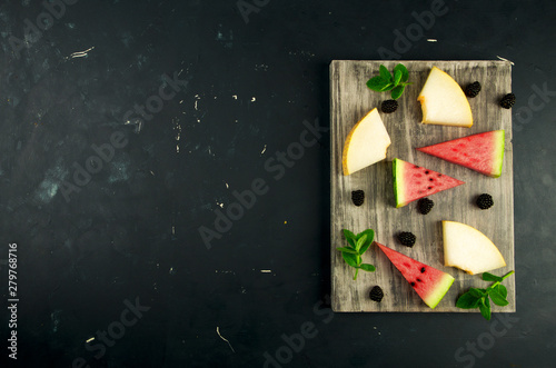 MELON WATERMELON BLACKBERRY AND MINT ON THE BACKGROUND. THREE PIECES OF RIPE WATERMELON JUICY MELON WITH RIPE BLACKBERRIES MINT LIE ON A WOODEN BOARD ON A DARK BACKGROUND.