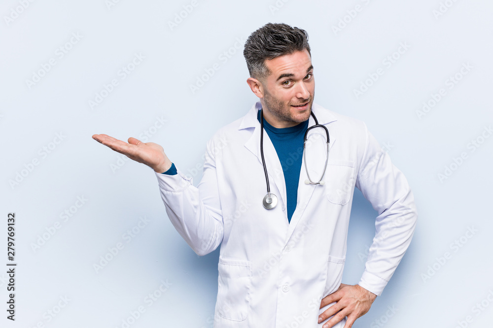 Young handsome doctor man showing a copy space on a palm and holding another hand on waist.