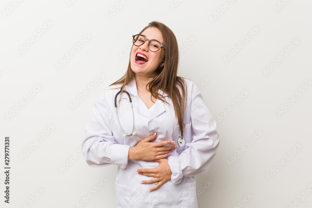 Young doctor woman against a white wall laughs happily and has fun keeping hands on stomach. Young doctor woman against a white wall laughs happily and has fun keeping hands on stomach.