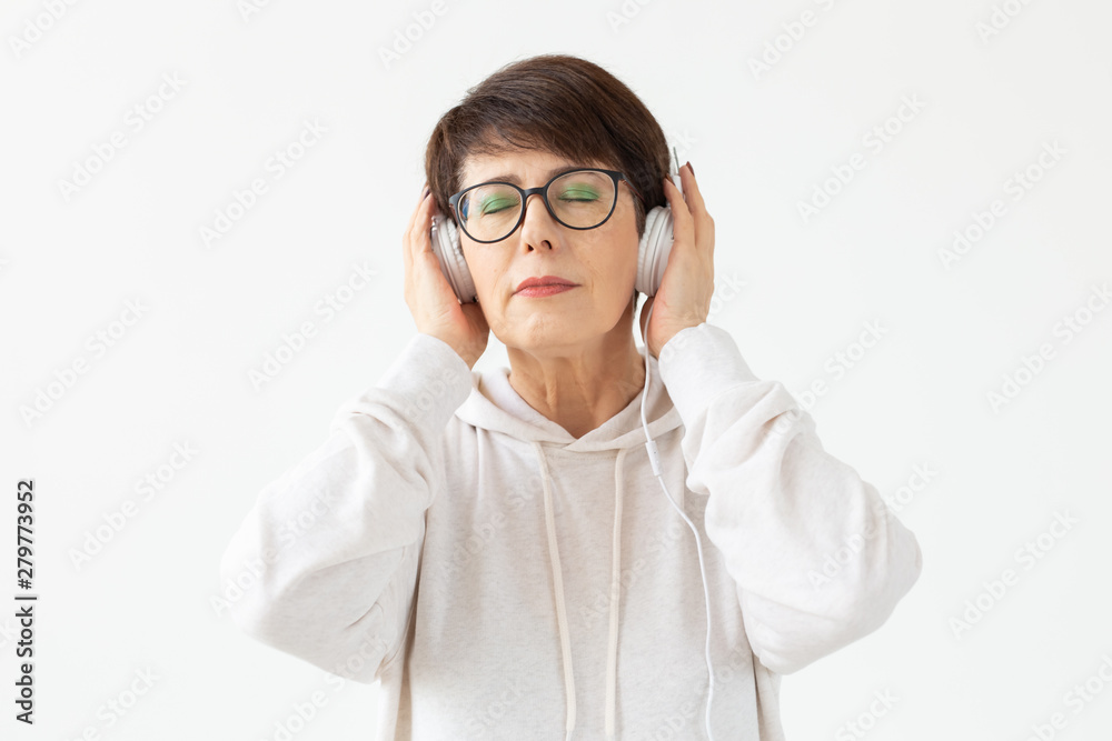 Cute positive middle-aged woman in sweater and glasses is listening to music with wire headphones standing on a white background. Concept of hobbies and subscriptions to favorite radio station. Cute positive middle-aged woman in sweater and glasses is listening to music with wire headphones standing on a white background. Concept of hobbies and subscriptions to favorite radio station.