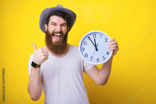 Handsome bearded gauy wearing a hat, excited holding clock and showing thumb up, over yelllow background