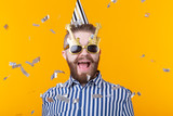 Joyful young hipster male in a paper cap and glasses is laughing happily among flying confetti on a yellow background. The concept of a mega party and holiday.