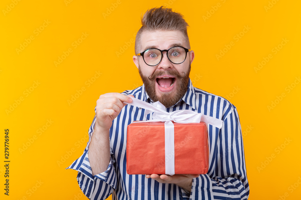 Surprised young hipster man with mustache and beard in surprise unpacks a red box with gifts on a yellow background, close up. The concept of gifts and surprises for the holiday Surprised young hipster man with mustache and beard in surprise unpacks a red box with gifts on a yellow background, close up. The concept of gifts and surprises for the holiday