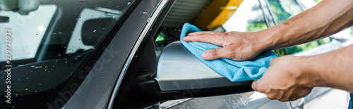 panoramic shot of man cleaning wet car mirror with blue rag