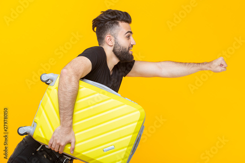 Cute pretty young arab man with a beard holding a yellow suitcase in his hands on a yellow background. Concept of travel and vacations