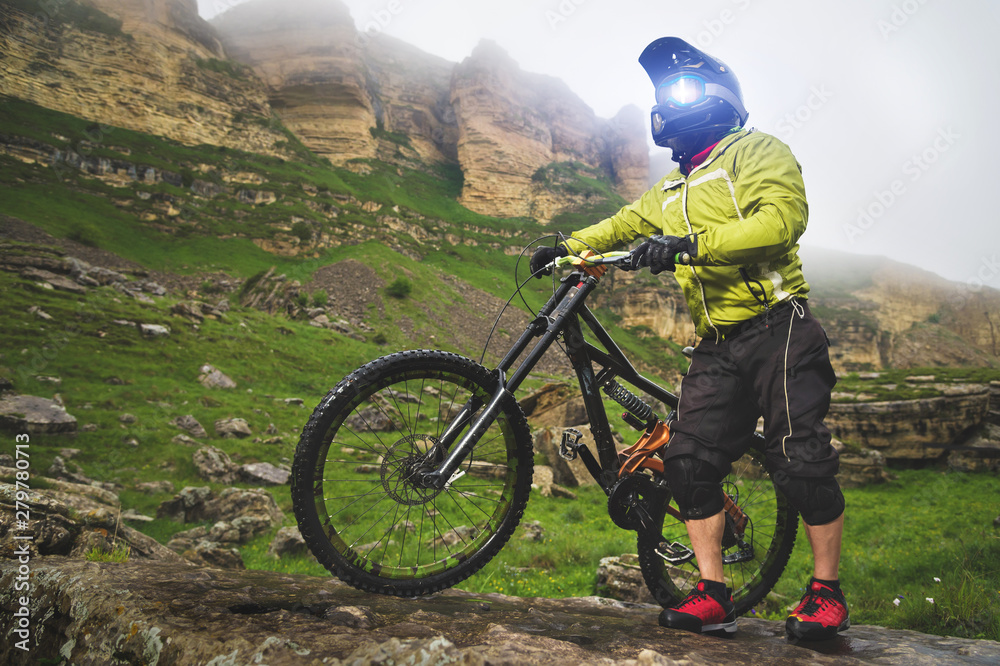 Naklejka premium Side view of a man on a mountain bike standing on a rocky terrain and looking at a rock. The concept of a mountain bike and mtb downhill