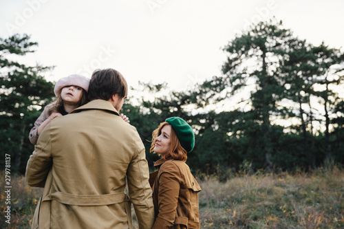 Young father, mother and little toddler daughter girl in a beret and a coat walk holding hands in the autumn forest.