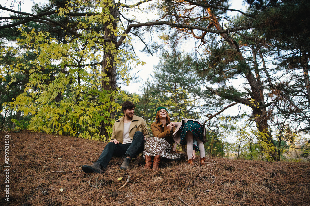 Young father, mother and little toddler daughter girl in a beret and a coat sitting on the ground in the autumn forest.