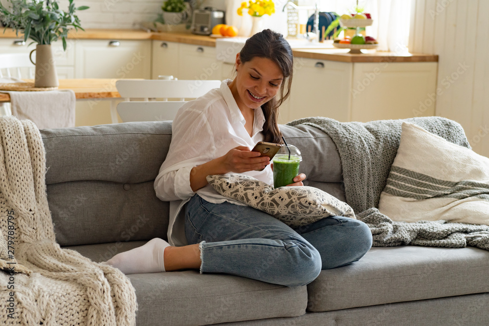 Young female drinking green smoothie sitting at the couch and looking at her smartphone at home