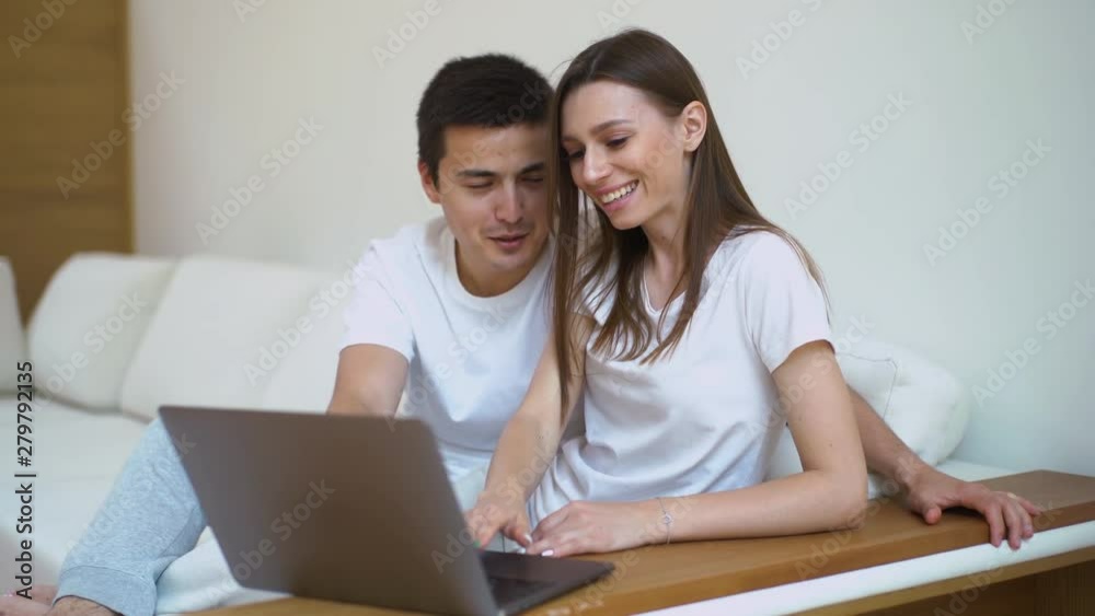 Young married couple sitting on soft sofa and looking at laptop in bright living room. Happy people buying things for house in online store sitting in light apartment. Guy and girl smiling. Young married couple sitting on soft sofa and looking at laptop in bright living room. Happy people buying things for house in online store sitting in light apartment. Guy and girl smiling.