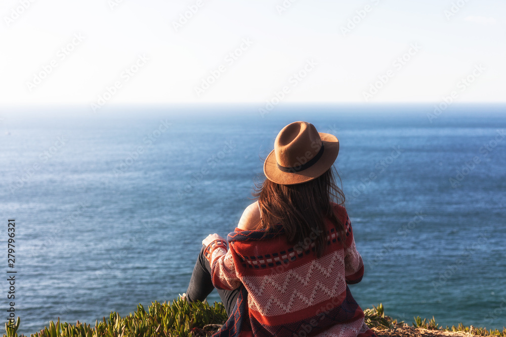 Closeup back view of woman in travel clothes and hat sitting and looking at blue ocean and sky.