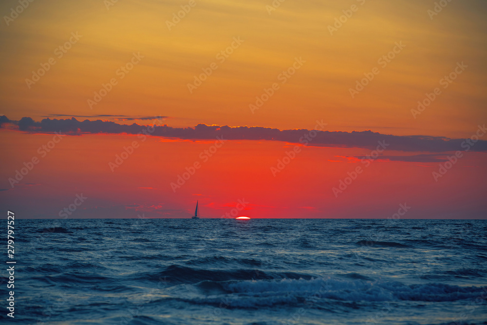 Sailboat on the horizon on the bright background of incredible sunset from the setting sun, the blue sea in the foreground