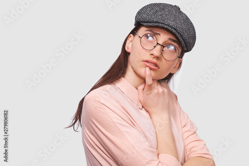 Thoughtful young woman wears transparent spectacles, casual pink shirt and gray cap, thinking and looking seriously at one side, posing against white studio background. People and emotions concept