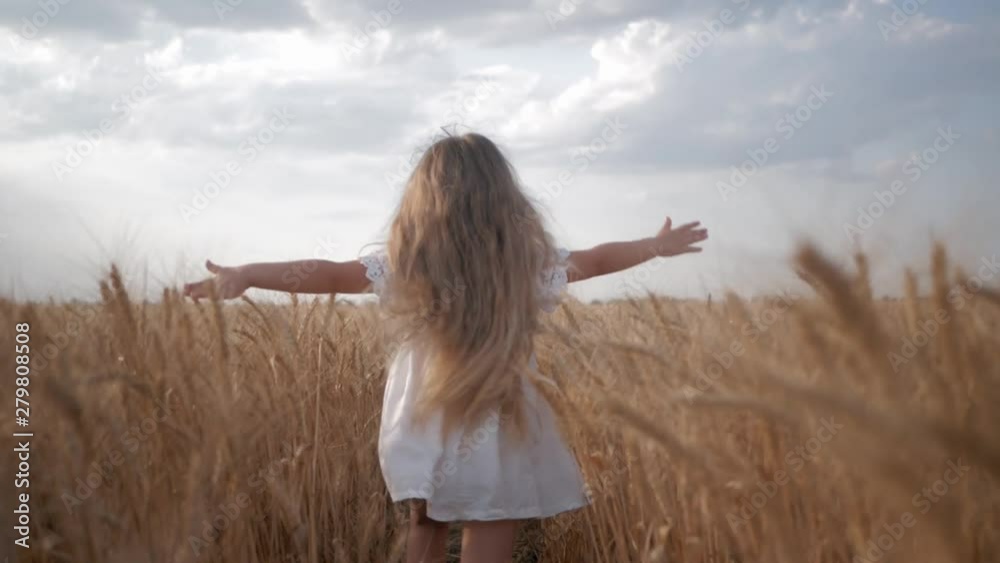happy childhood, little kid girl in white dress runs with arms spread to sides across wheat field with golden spikelets of grain in autumn yield season against blue sky
