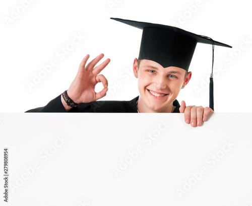 Male graduate student peeking from behind a blank panel, isolated on white background. Handsome graduate guy student in mantle showing blank placard board and making Ok sign. photo