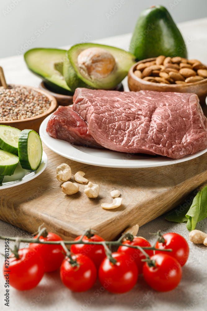 raw meat on wooden chopping board near nuts and vegetables isolated on grey, ketogenic diet menu