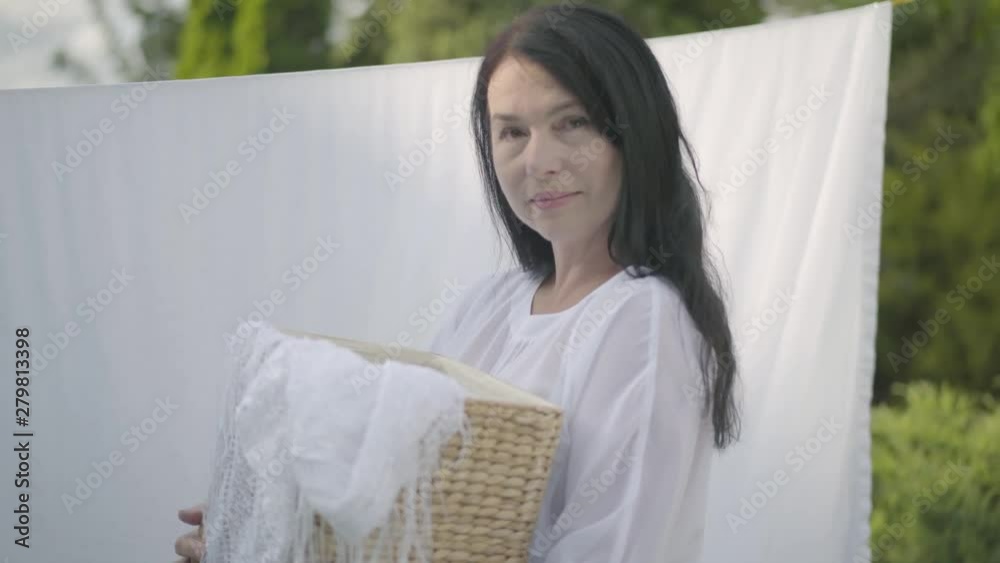 Adorable mature woman with long black hair holding wicker basket in hands while hanging white clothes on a clothesline outdoors and looking at camera. Washday. Senior woman doing laundry. Adorable mature woman with long black hair holding wicker basket in hands while hanging white clothes on a clothesline outdoors and looking at camera. Washday. Senior woman doing laundry.