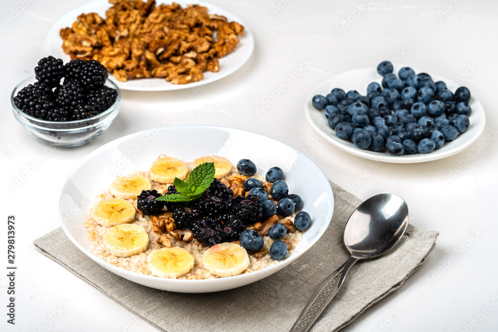 Oatmeal porridge with bananas, walnuts, blackberries, blueberries, honey and mint in a white bowl on a white background. Healthy breakfast and homemade diet food. Oatmeal porridge with bananas, walnuts, blackberries, blueberries, honey and mint in a white bowl on a white background. Healthy breakfast and homemade diet food.