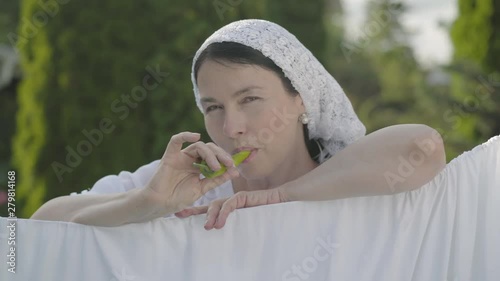 Attractive senior woman with shawl on her head eating green pepper looking at camera smiling over the clothesline outdoors. Washday. Pretty housewife doing laundry. photo