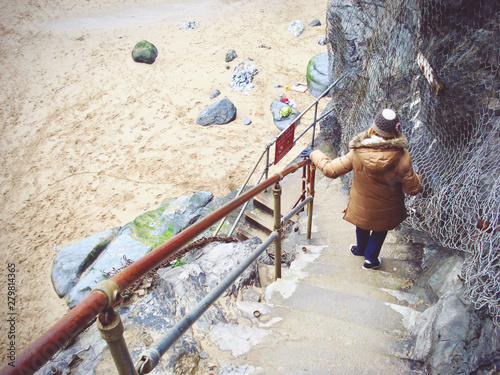 Young woman in winter puff jacket and woolen hat holding the fence and descending concrete stairs towards the sandy beach, in Bedruthan Steps Beach, Cornwall, UK