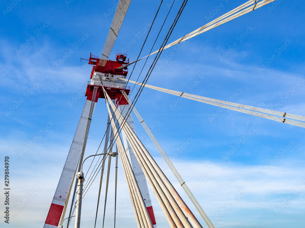 Cable-stayed bridge with high red and white tower and steel cables against blue sky. Cable-stayed bridge with high red and white tower and steel cables against blue sky.