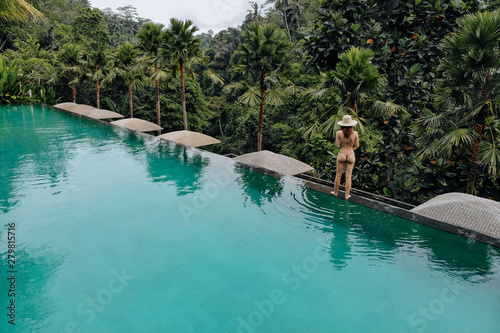 Woman walking on edge of infinity pool in beige bikini and straw hat in jungle around trees. Vacation concept