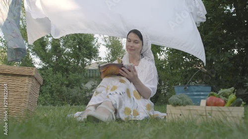 Adorable woman with a white shawl on her head resting in the garden sitting on the grass in front of clothesline reading magazine. Positive housewife resting after doing laundry. Bottom view photo