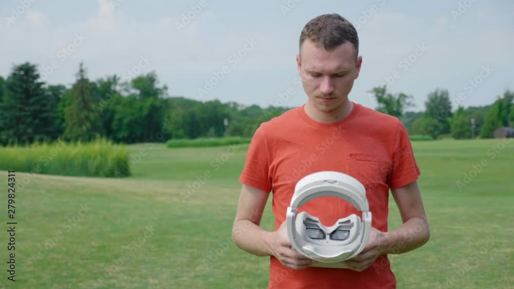 A man holds in hands a VR headset in the park. A young guy in a red T-shirt and a helmet is experiencing virtual reality against the background of nature. A human on the right in the frame. 4K footage A man holds in hands a VR headset in the park. A young guy in a red T-shirt and a helmet is experiencing virtual reality against the background of nature. A human on the right in the frame. 4K footage