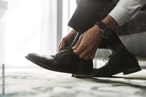 Cropped image closeup of successful man tying his shoe laces while sitting on bed in hotel room during business trip