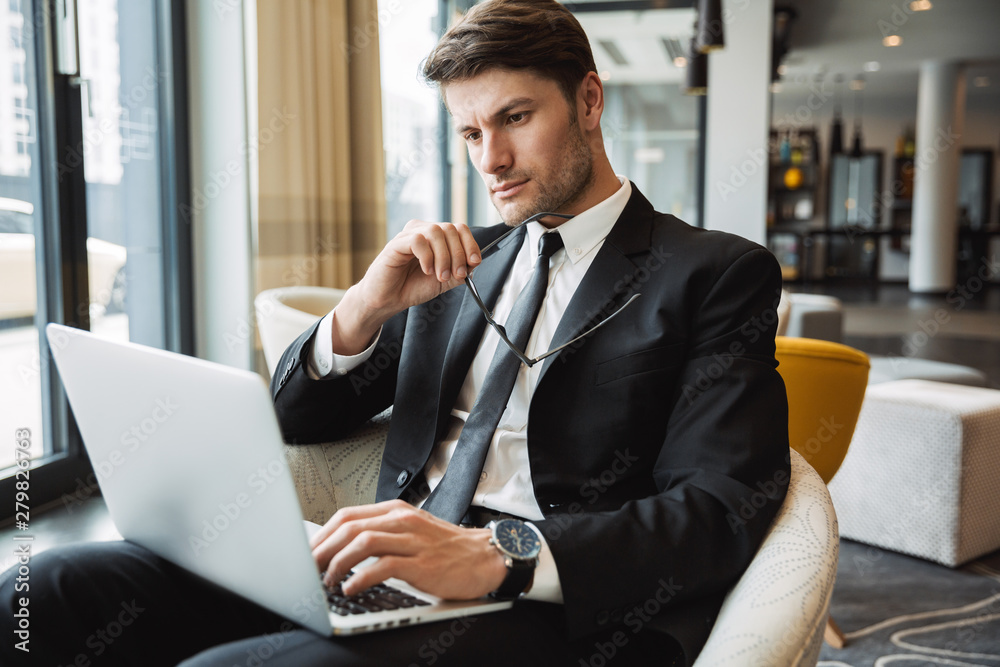Portrait of serious young businessman sitting on armchair with laptop computer in hotel hall Portrait of serious young businessman sitting on armchair with laptop computer in hotel hall