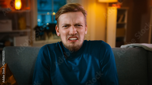 Portrait of a Handsome Man Sitting on a Couch at Home at Night, Watching Gore Horror Movie on TV. He Gets Really Scared, Confused and Disgusted.