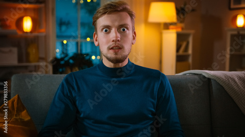 Portrait of a Handsome Man Sitting on a Couch at Home at Night, Watching Entertaining Movie on TV. He is Really Mind Blown and Shocked by Huge Sales and Discounts Showed in Advertisement. photo