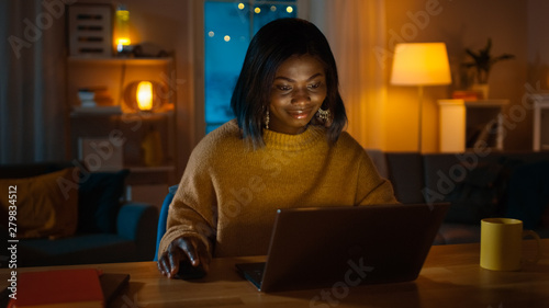 Portrait of Beautiful Smiling African American Girl Using Laptop while Sitting at Her Desk at Home. In the Evening Creative Woman Works on a Computer In Her Cozy Living Room. 